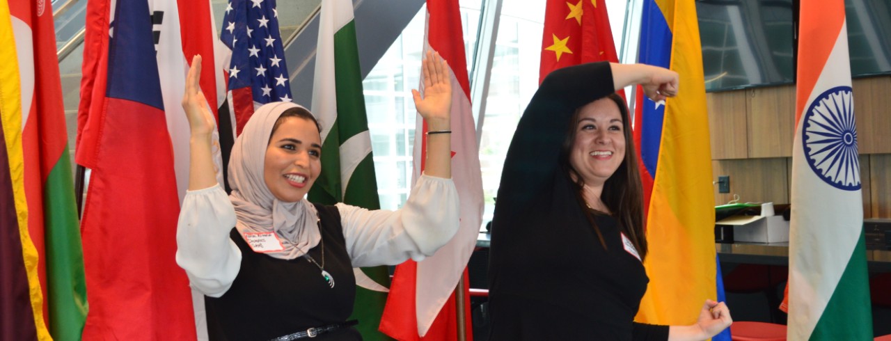 Two women form the letters UC with their arms, standing in front of flags from a variety of countries.