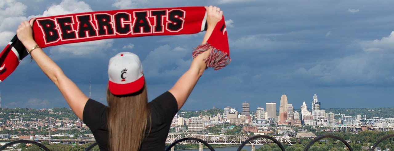 Female wearing UC hat, holding up Bearcats scarf with city skyline in background.