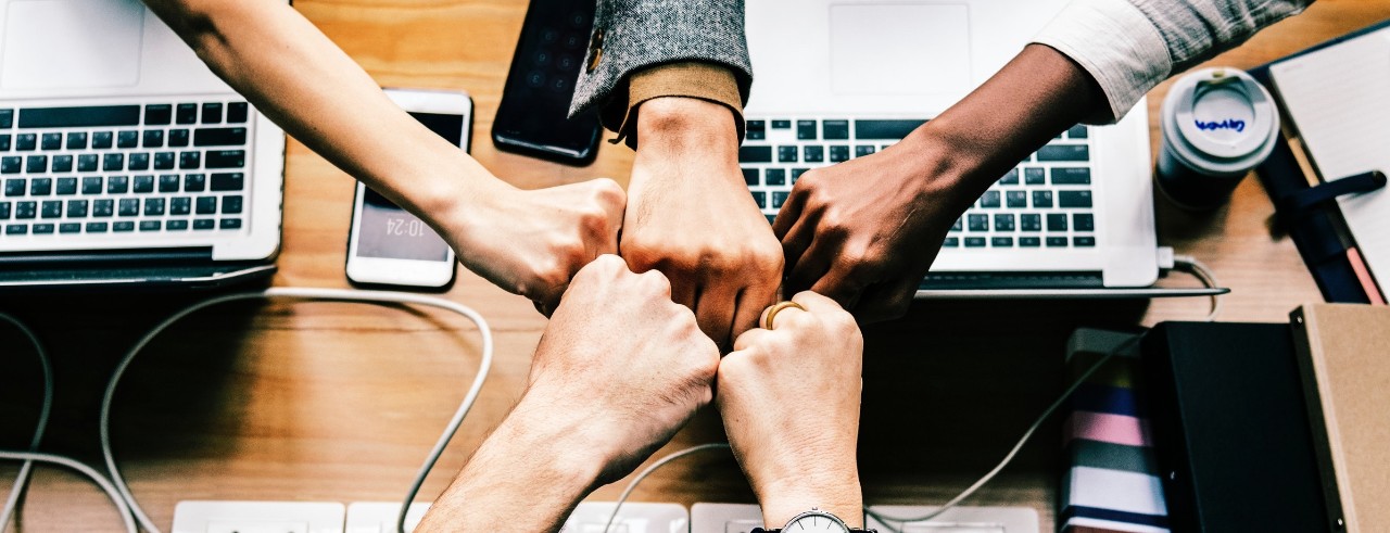 Desk with several open computers; five hands bumping fists together in teamwork