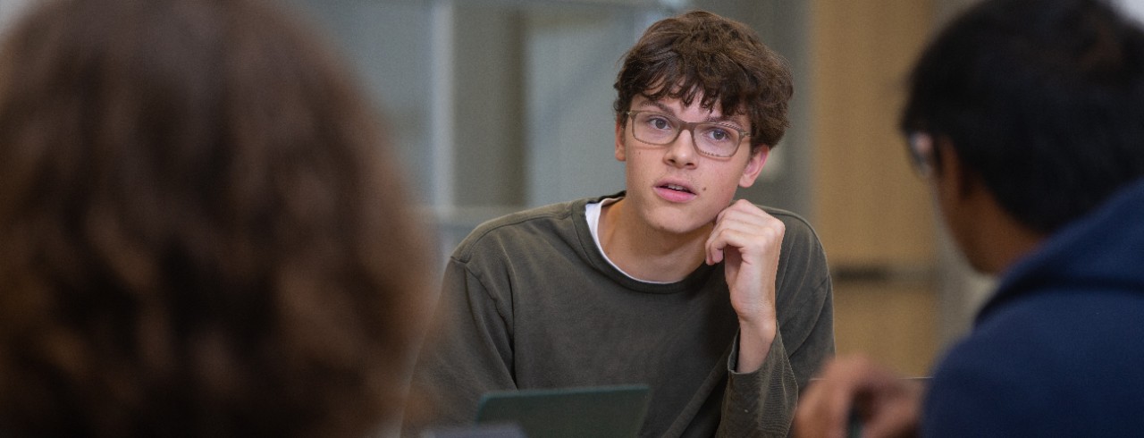 A UC student seated at a table engaged in conversation with other students.