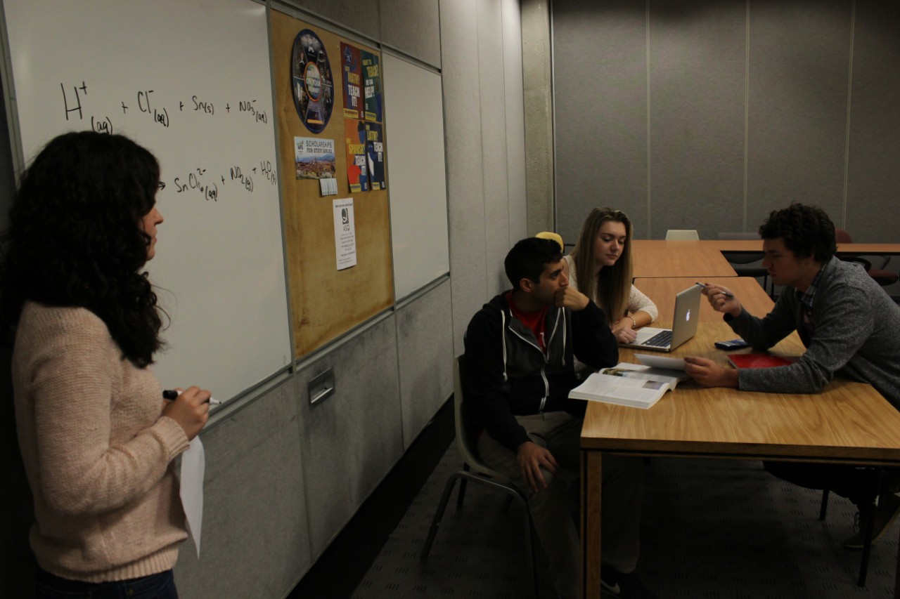 A group of University of Cincinnati students looking up at a student writing on a whiteboard in the Learning Commons