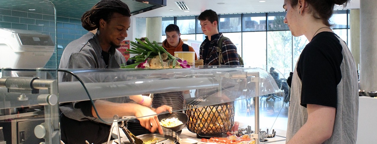 Three male students at serving stationg in dining center On the Green. A food service worker creates a made-to-order bowl.