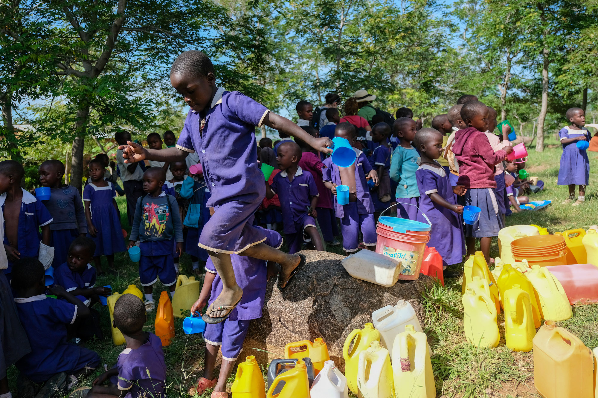 Tanzanian child jumps over plastic pitchers with dozens of kids in background