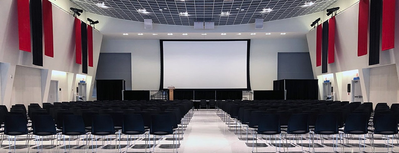 Round tables with chairs and banners on the wall in great hall. A stage with a podium and screen are at the end of the great hall.