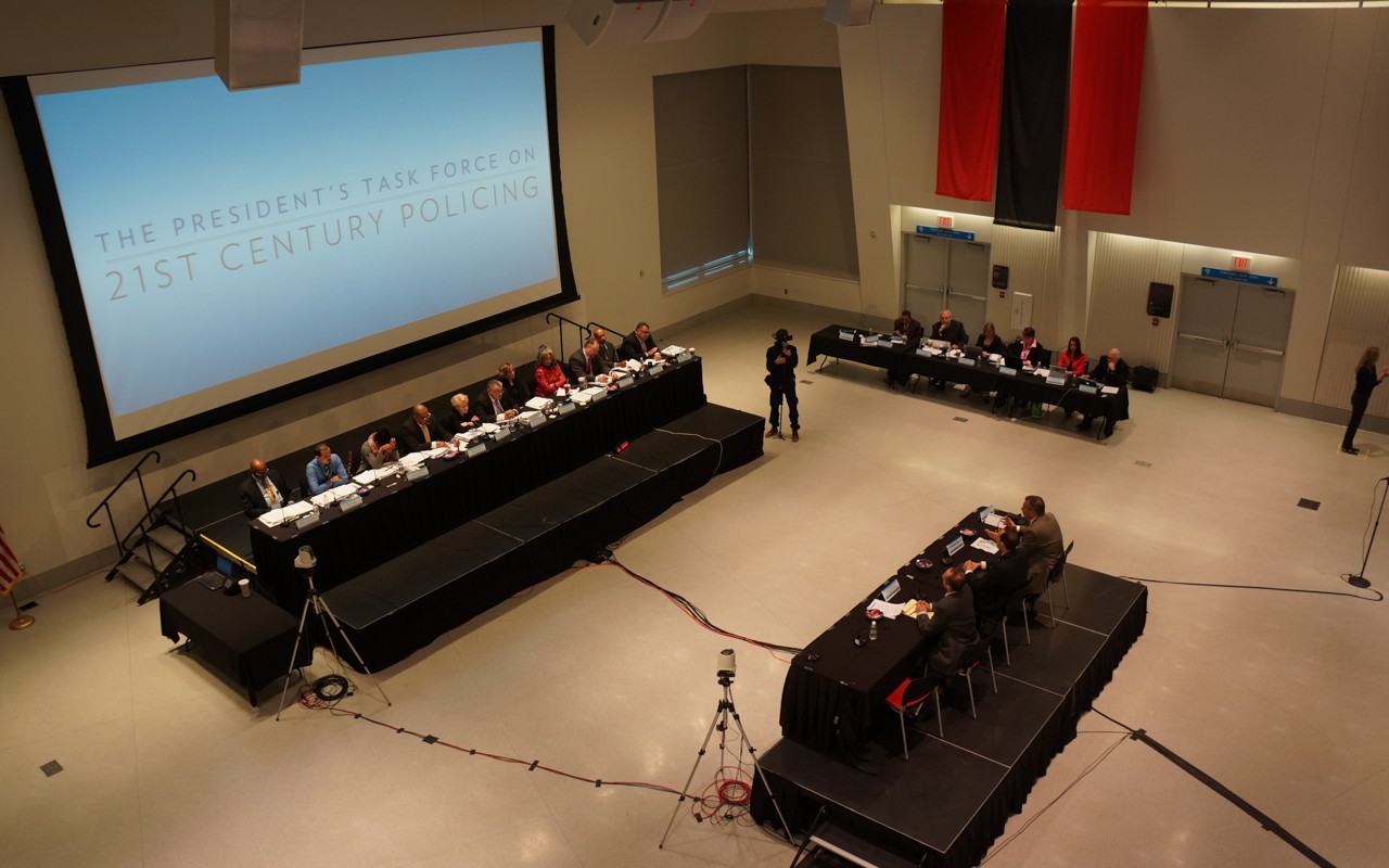 A large meeting taking place with a long table on the stage with several presenters sitting at it. There is another smaller stage across from the main one that also has several attendees sitting at a rectangular table. There is a third table to the side of the main table with attendees sitting at the table.
