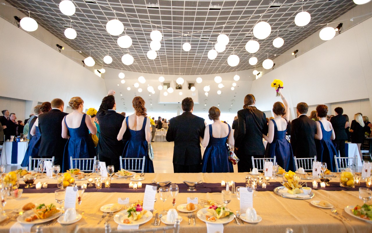 A wedding party standing with their back to their dinner table. The women are dressed in navy gowns while the men are wearing formal black suits. The table is covered with a gold table cloth and matching dinnerware. 