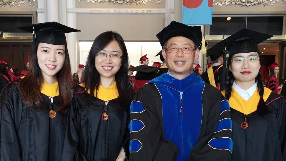 International faculty member celebrates with three students in graduation gowns.