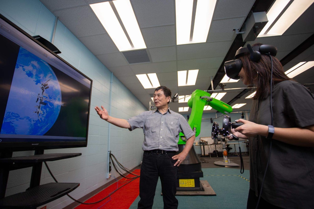 A professor gestures at a screen while a student wears a VR headset