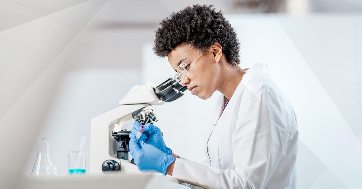 A black woman with short black hair wears a white lab coat and blue surgical gloves. She looks into a microscope atop a white counter.