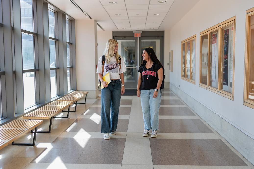 Two UC students walking down a naturally-lit hallway, discussing what college classes are like