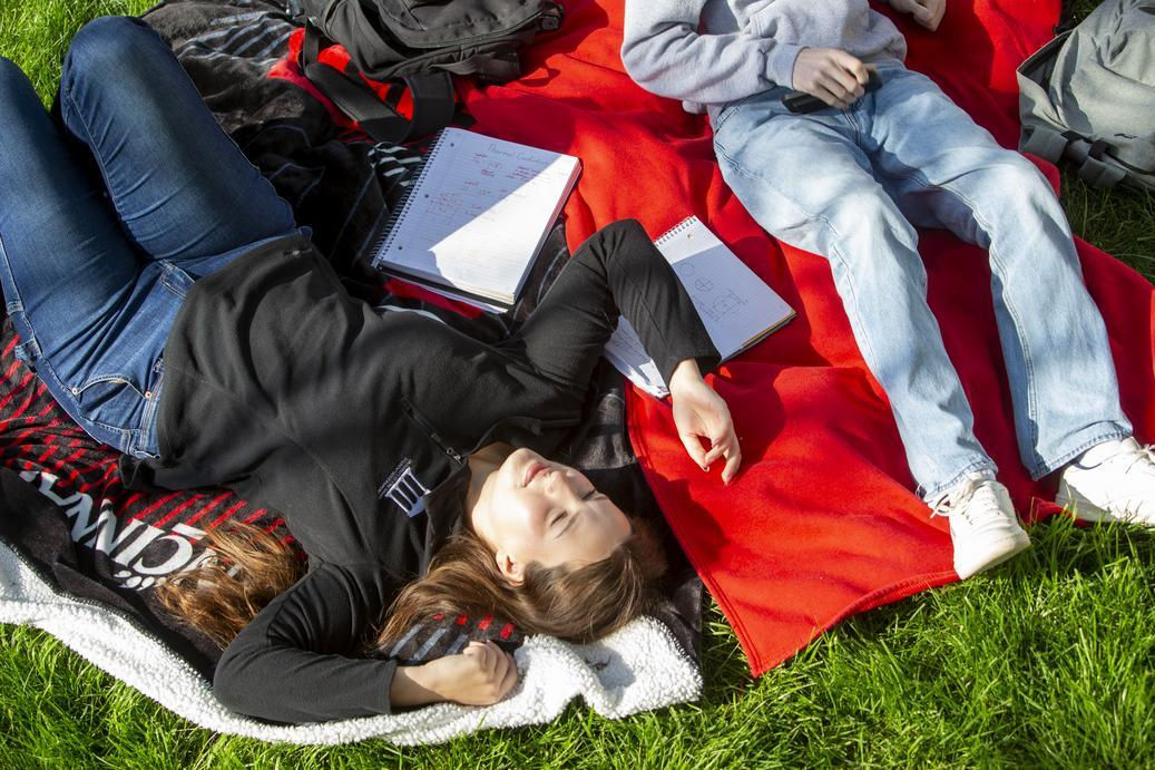 UC students laying on a blanket on campus green as they talk about if it's cheaper to live on campus or off campus