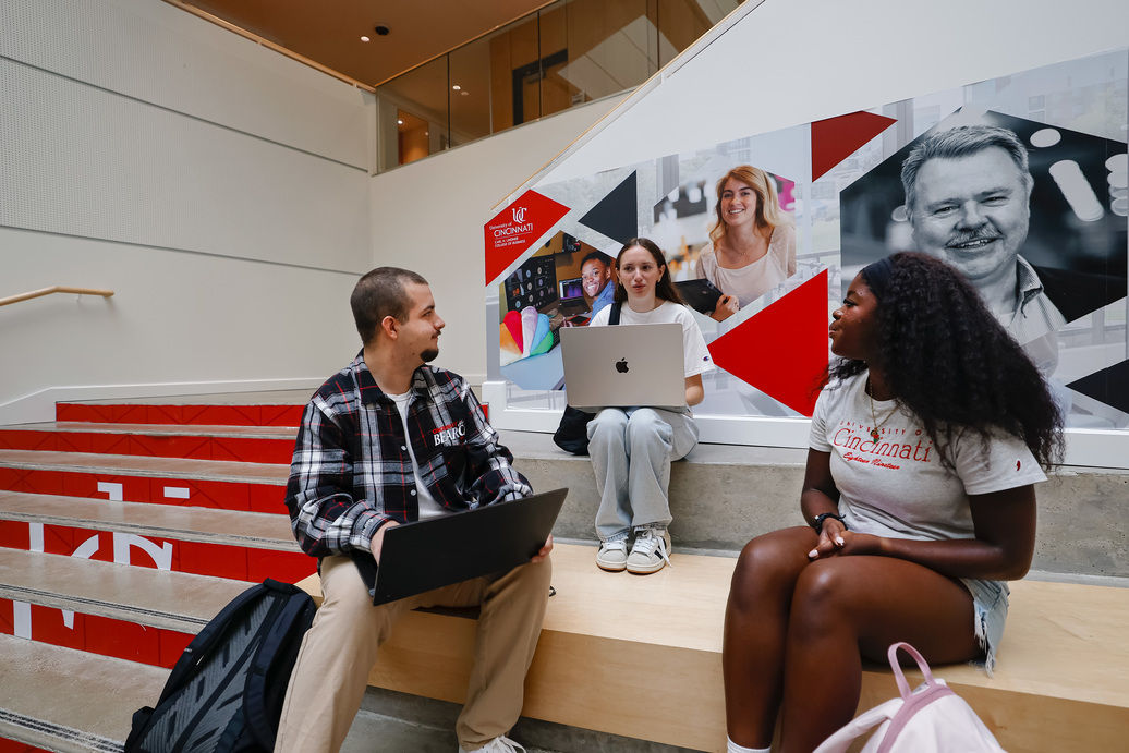 Three students sit on steps in a UC building talking about public vs private college.