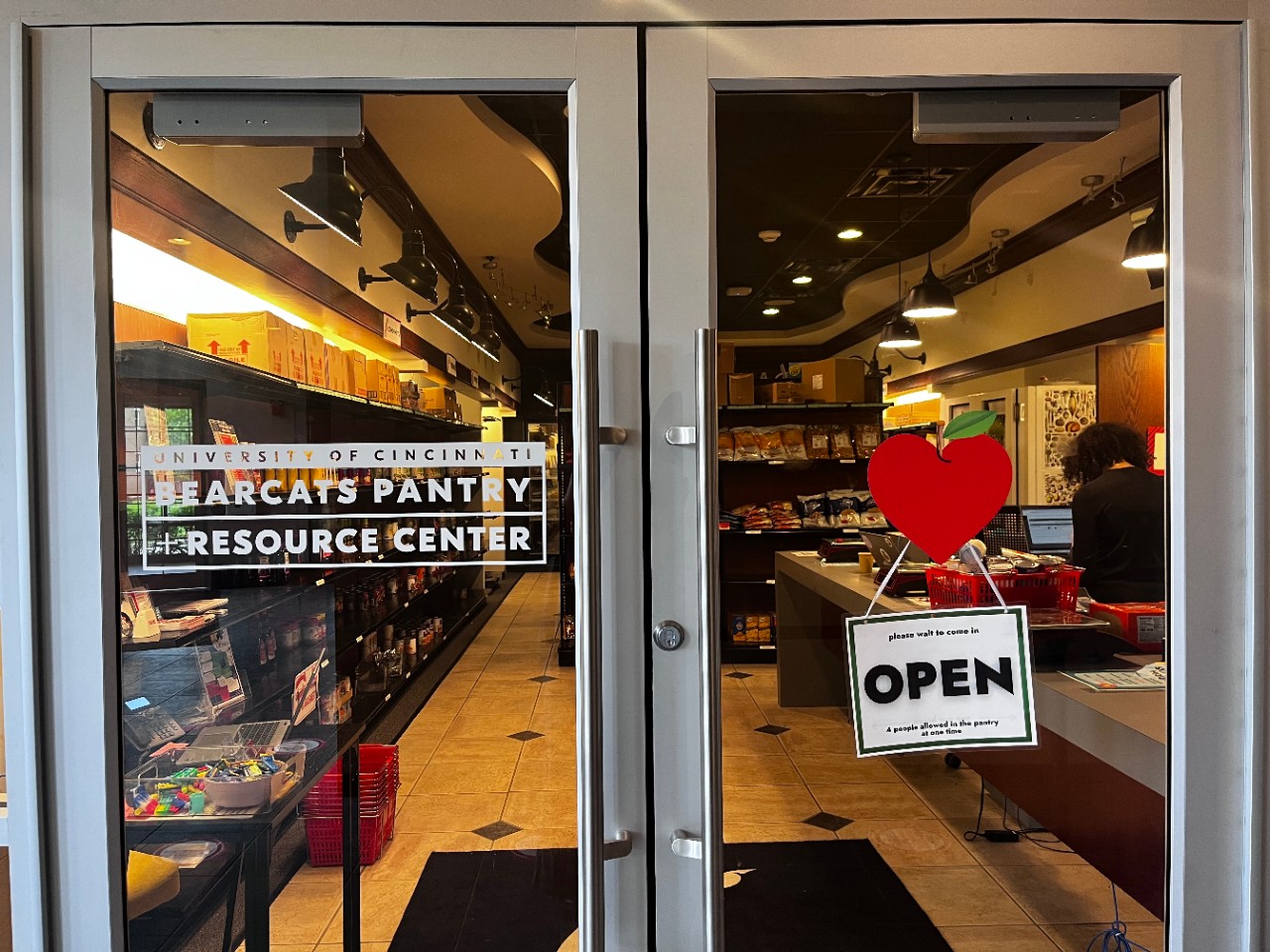 Looking through clear glass entry doors into the Bearcats Pantry where there are shelves of food. A sign on the door reads "University of Cincinnati Bearcats Pantry and Resource Center" and "Open."