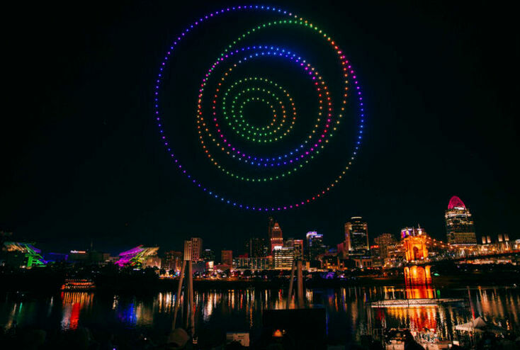 A colorful Cincinnati skyline at night with glowing multicolored drone lights creating a circular pattern in the sky.