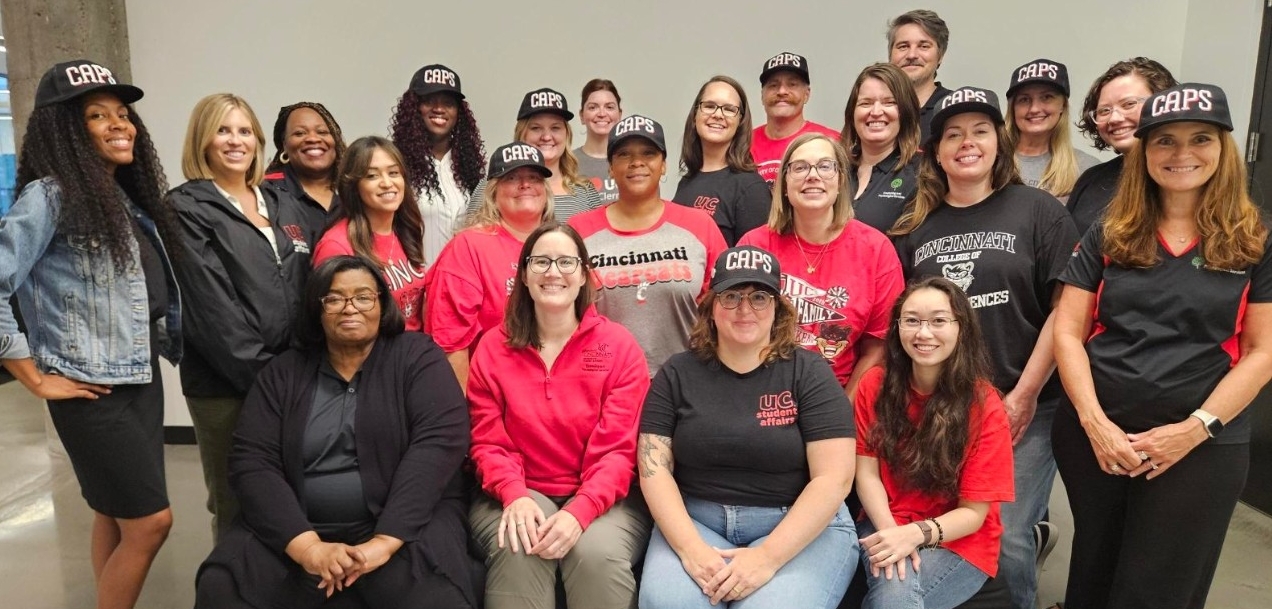 Group picture of CAPS staff. Some individuals are sitting and the majority of the group is standing, wear black and red attire and our CAPS baseball hats. 