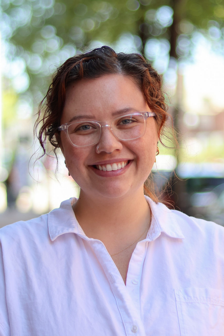 Photograph of Aislinn Burns outside of the CAPS building. She is wearing a white top. and glasses 