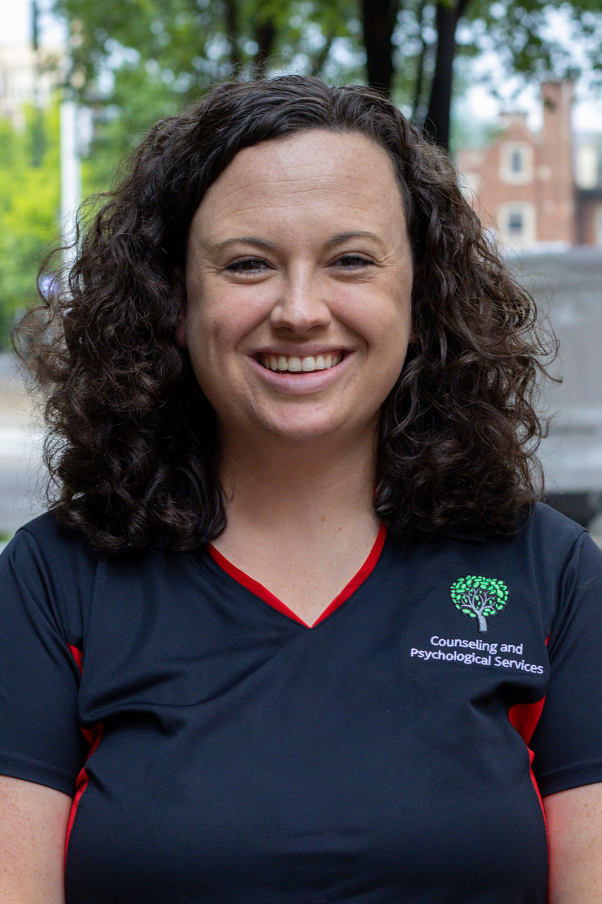 Photograph of Angela Hinrichsen smiling outside CAPS building. She is wearing a CAPS polo. 