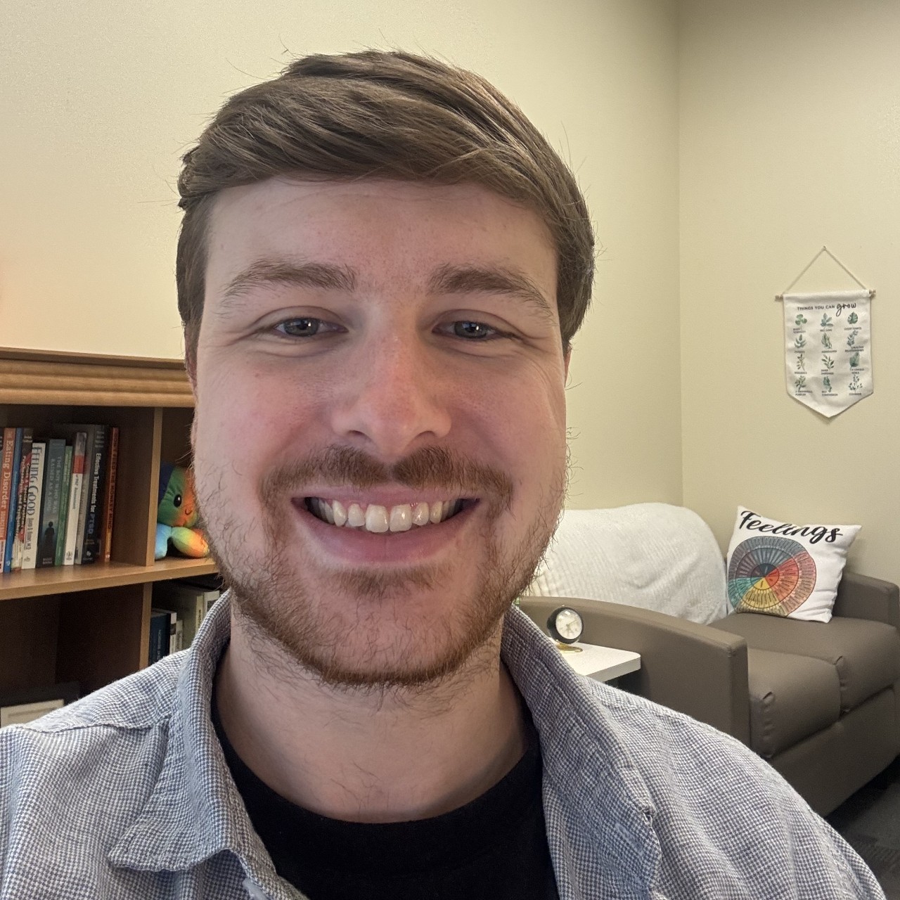 Photograph of Austin Nader smiling in his office. He is wearing a gray button up over a black shirt. 