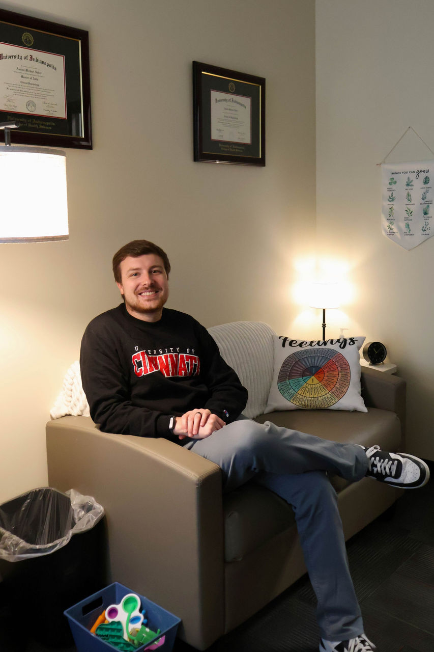 Photograph of Austin Nader smiling in his  office. He is wearing a UC sweatshirt. 