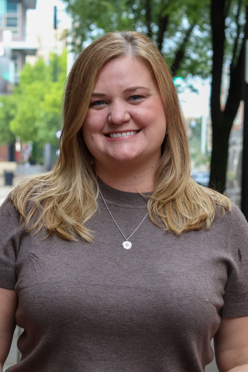 Photograph of Caity Cavanaugh smiling outside of CAPS office. She is wearing a tan top. 