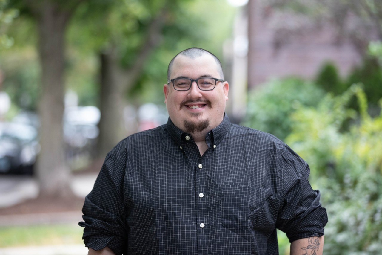 Photograph of Christopher Nora smiling as he stands outside. He is wearing glasses and a striped button up shirt. 