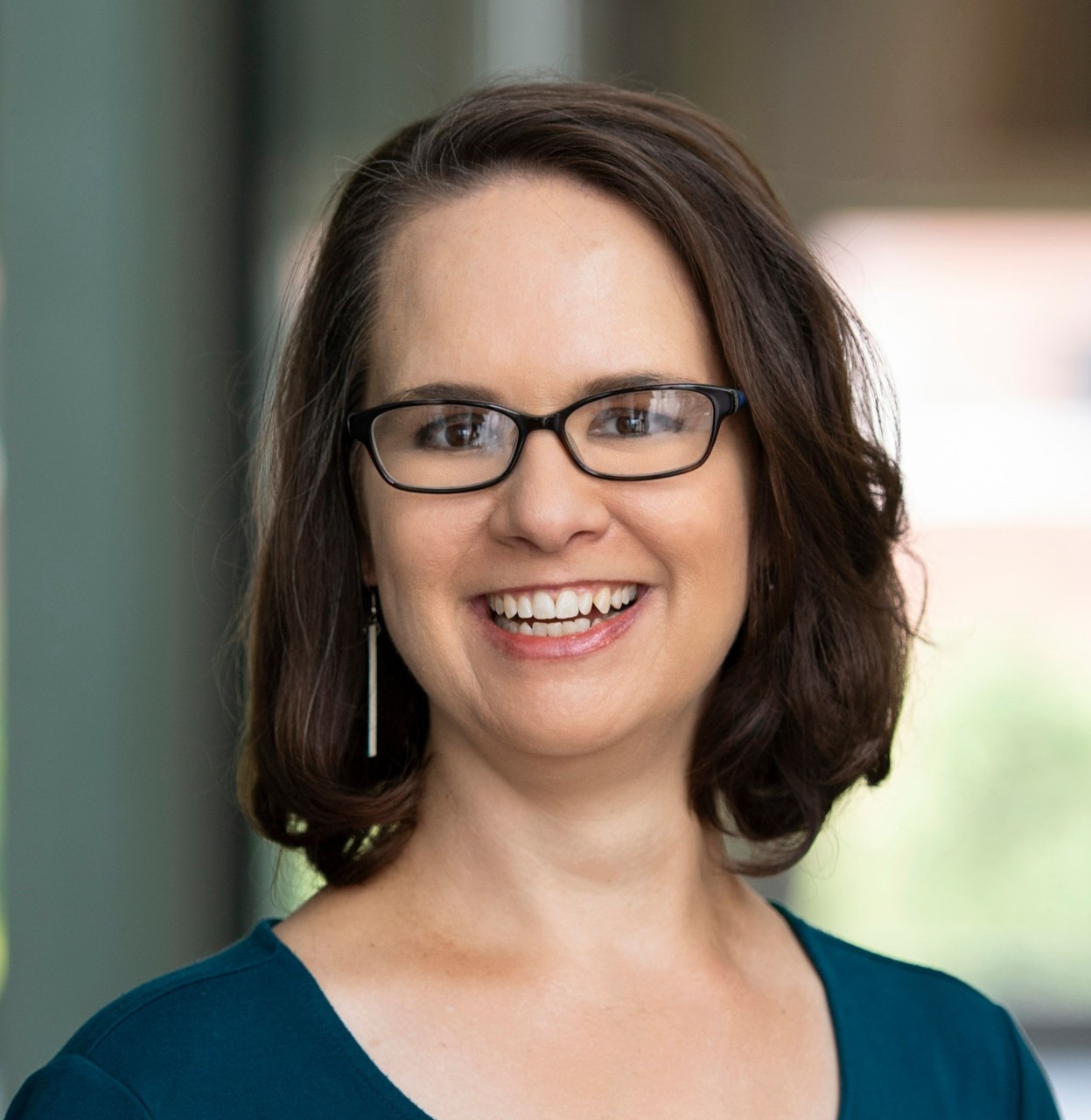 Photograph of Christy Wolfram smiling in a professional, outdoor headshot. She is wearing glasses and a green top. 