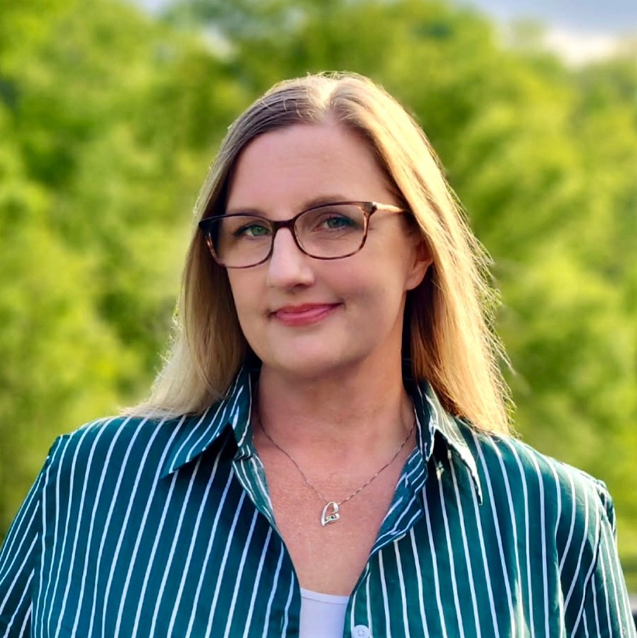Photograph of Danielle Drake smiling outside on a sunny day. She is wearing glasses and a blue striped shirt.