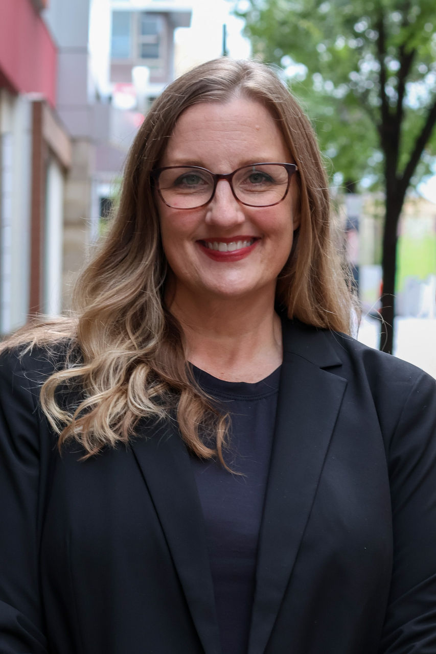 Photograph of Danielle Drake smiling outside of CAPS building. She is wearing glasses and black top and black dress jacket.