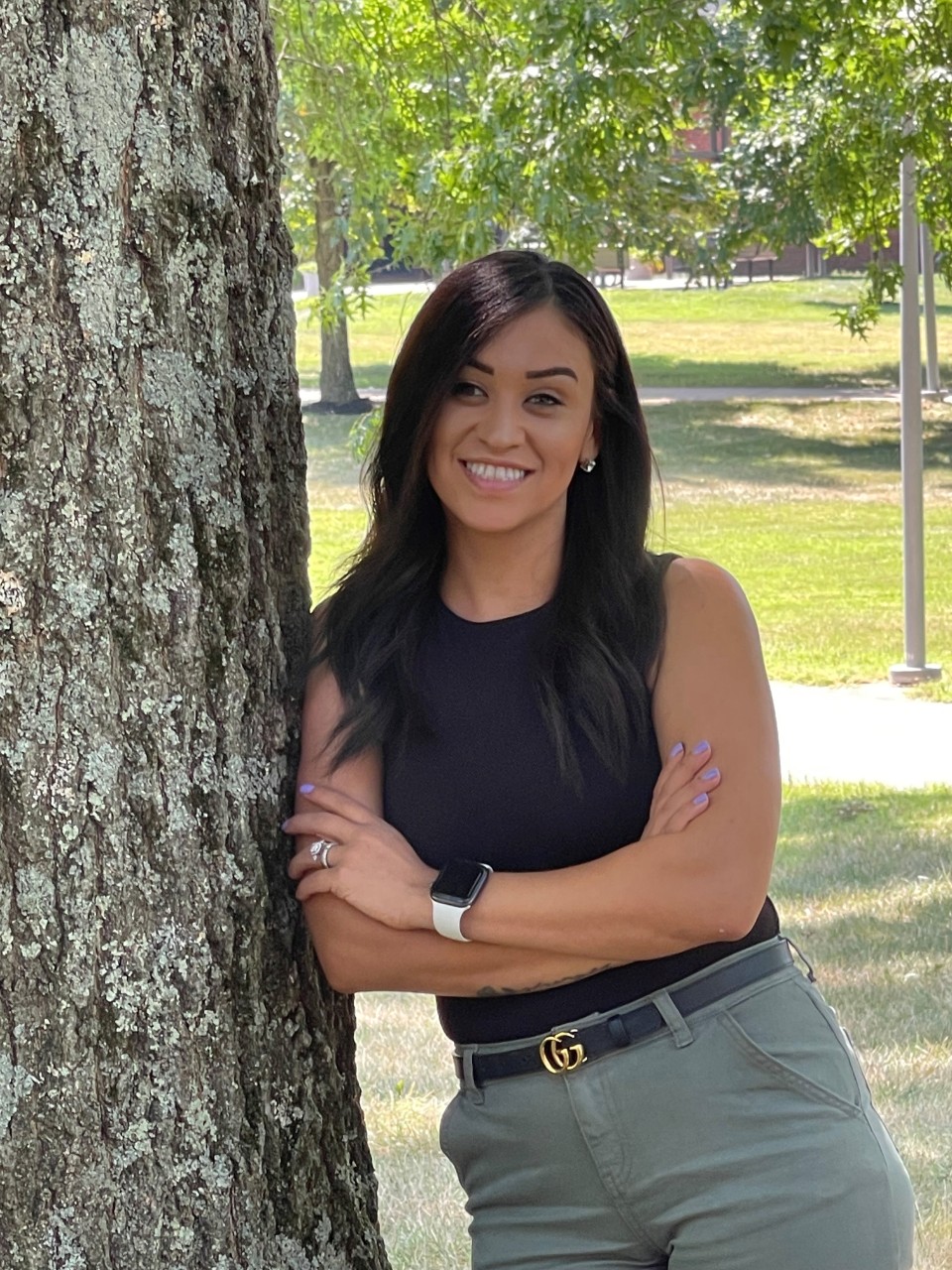 Photograph of Desiree Gonzales standing outside and leaning against a tree. She is smiling and wearing a black top.