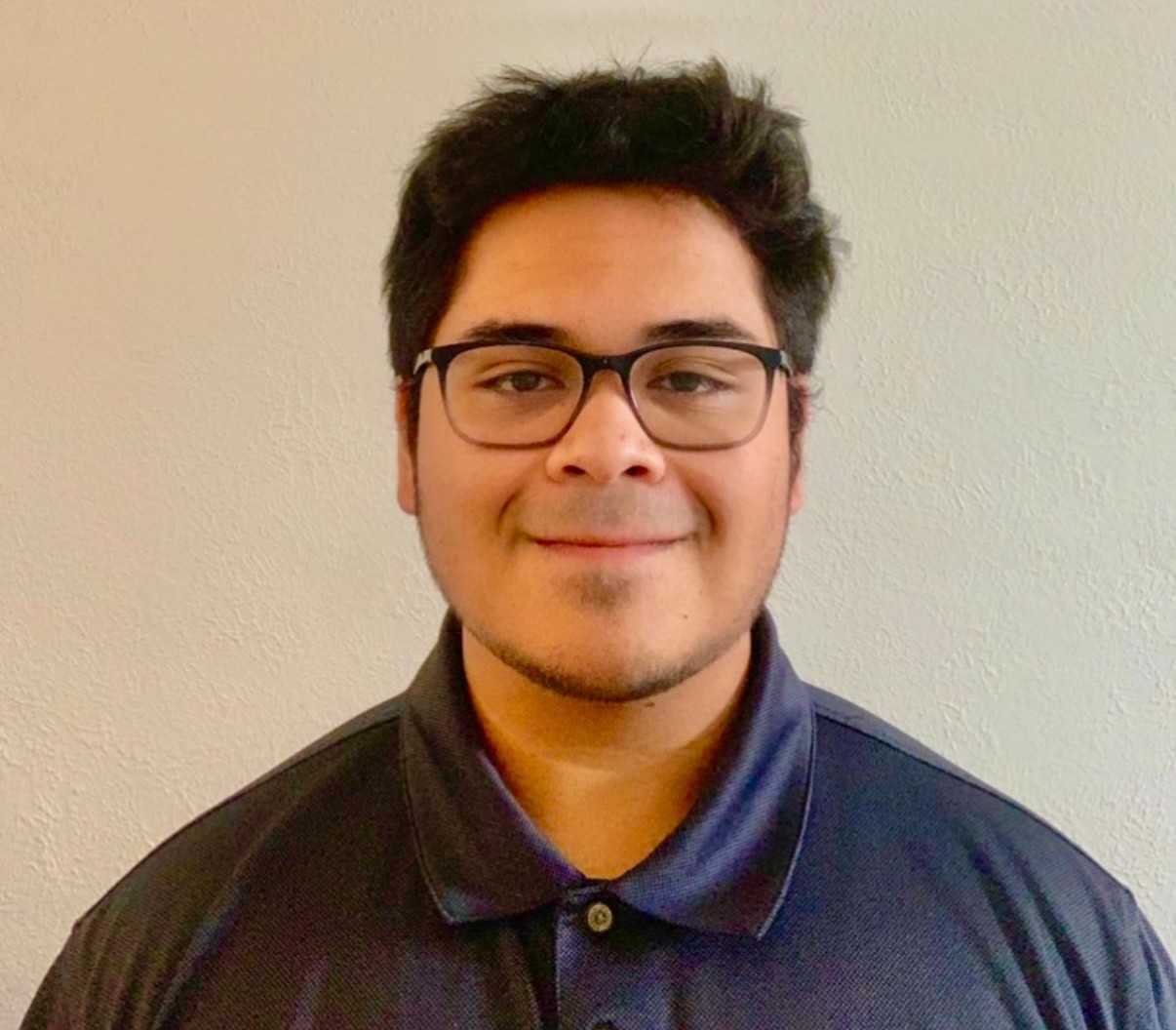 Photograph of Grayling Keune smiling in front of a light yellow wall. He is wearing glasses and a blue polo. 