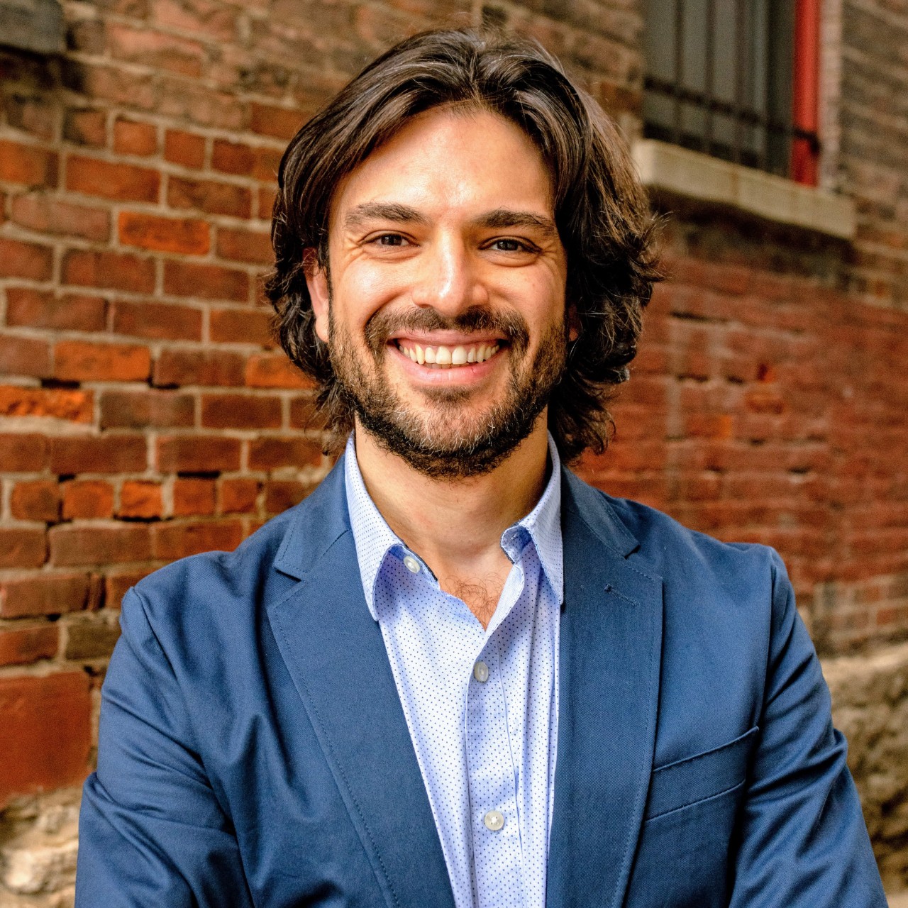 Photograph of Gianluca Bruno standing outside with a brick wall behind him. He is smiling and wearing blue.