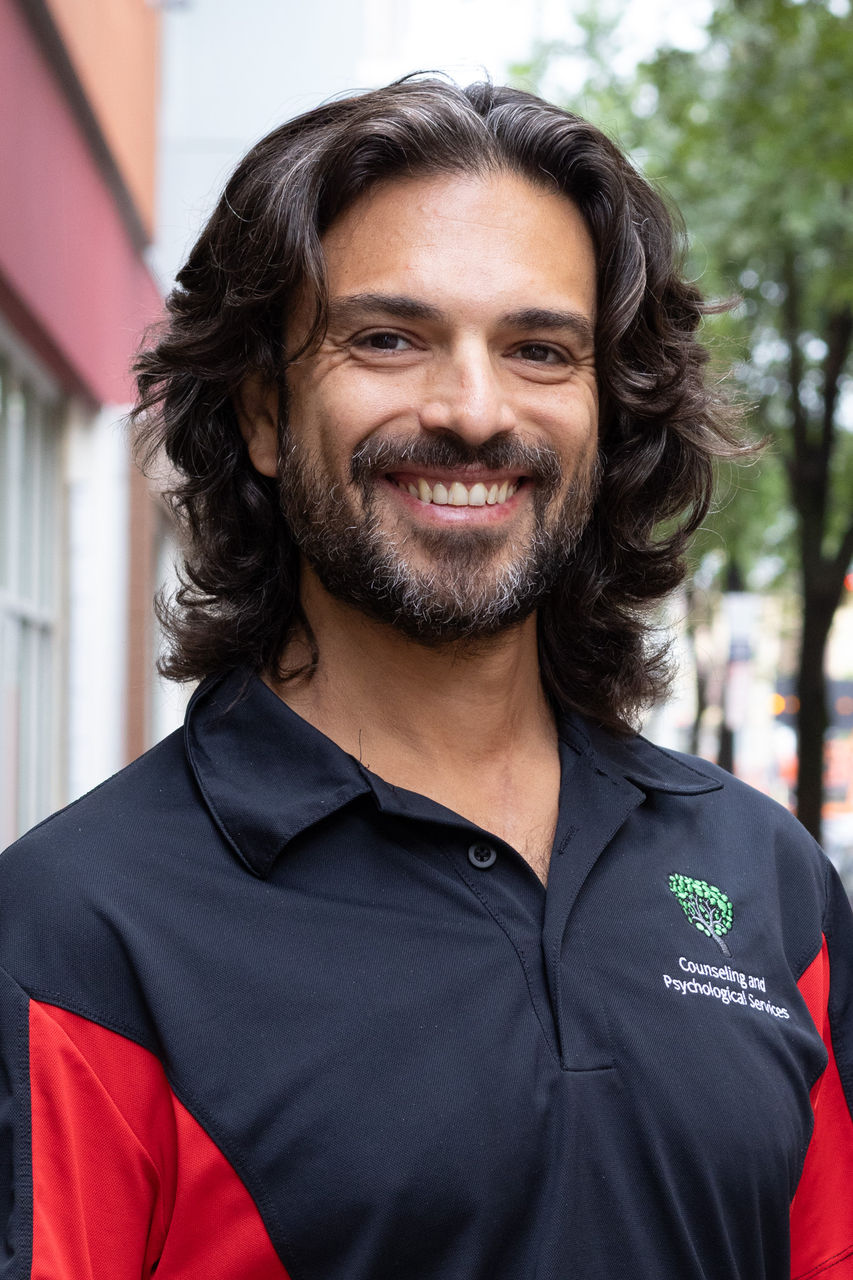 Photograph of Gianluca Bruno standing outside of CAPS building. He is smiling and wearing CAPS polo.