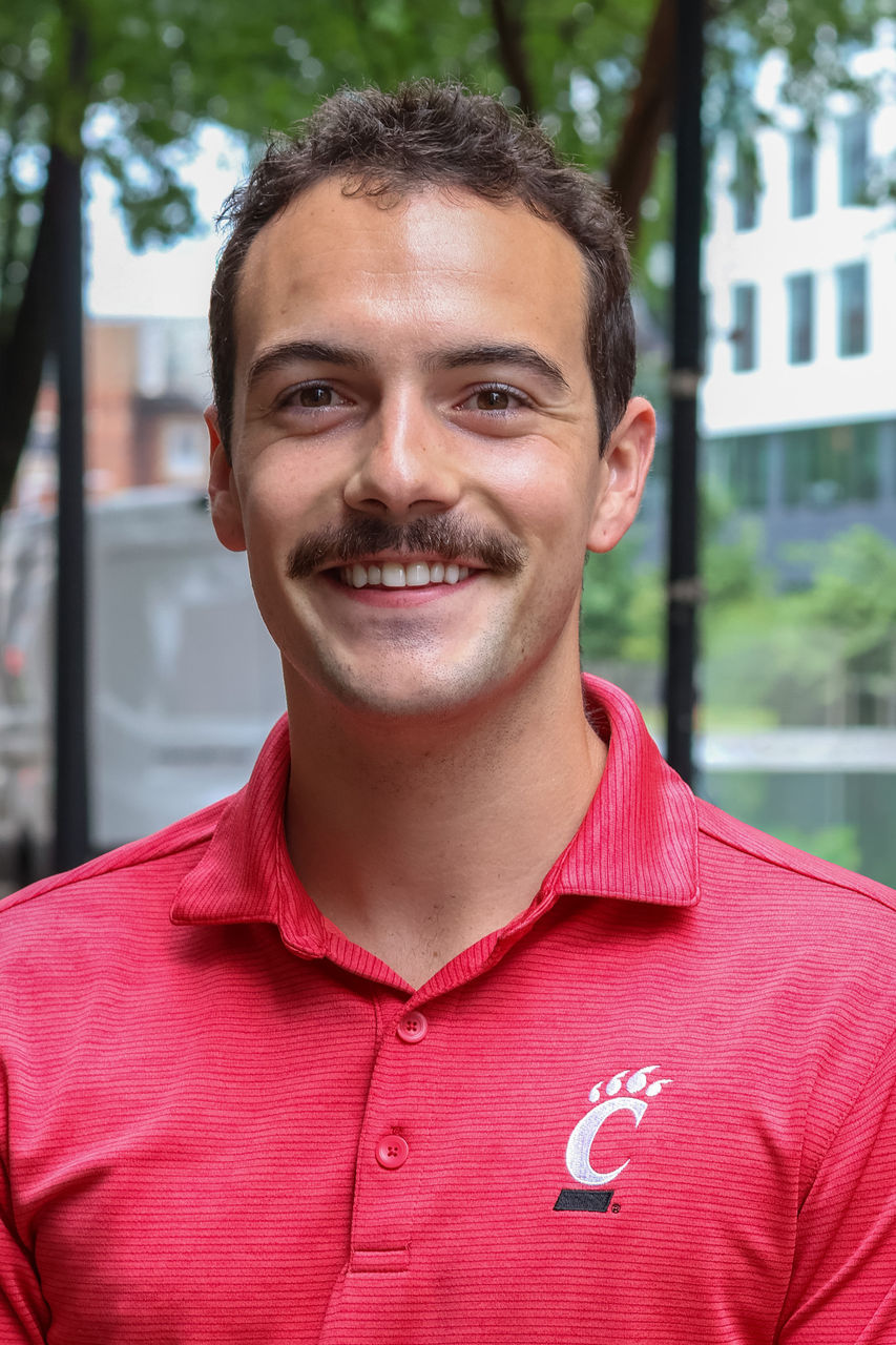 Photograph of Harry Powers smiling outside of CAPS building. He is wearing a red UC polo.
