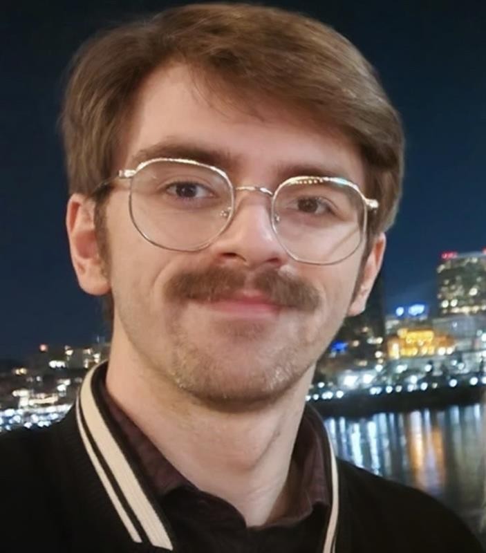 Photograph of Kobe Lyons smiling with the skyline behind him at night. He is wearing glasses and a jacket. 