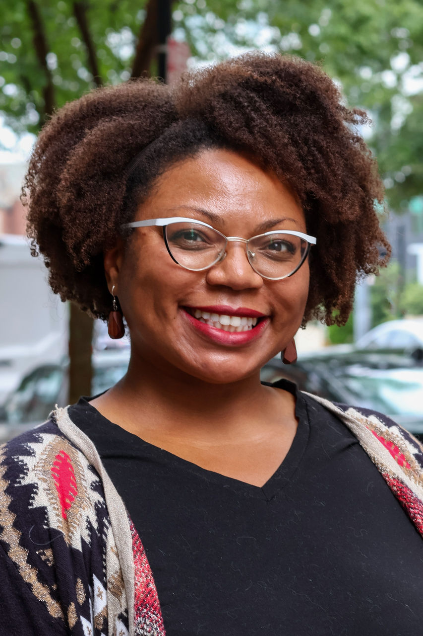 Photograph of Lianna Tullis-Robinson smiling outside of the CAPS building. She is wearing glasses and a red/black/tan patterned sweater.
