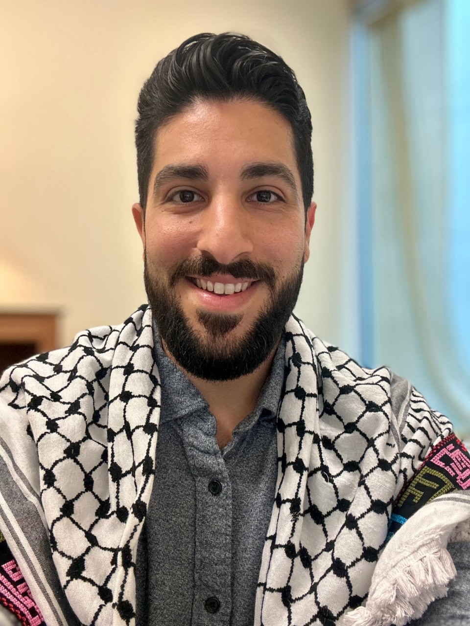 Photograph of Mike Madanat smiling in his office. He is wearing a keffiyeh.