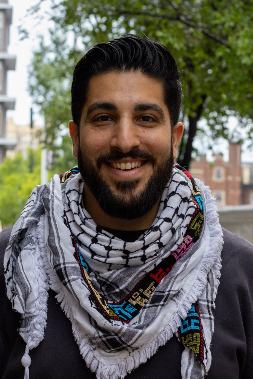 Photograph of Mike smiling outside of CAPS building. He is wearing a keffiyeh.