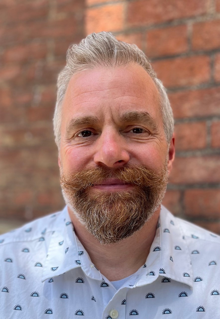 Photograph of Pete Mesrin smiling, standing outside in front of brick wall. He is wearing a white button up shirt with patterned half-suns.