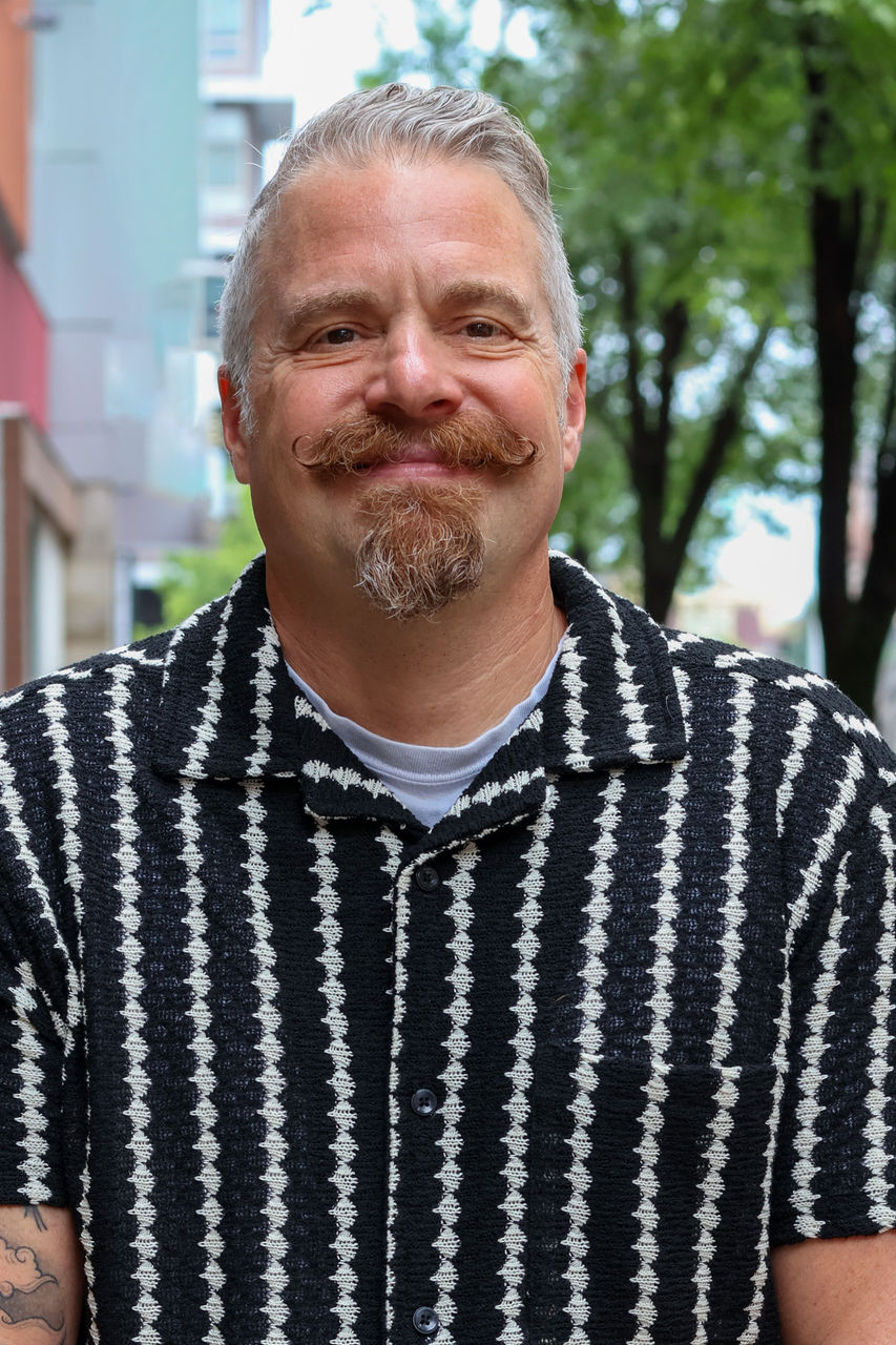 Photograph of Pete Mesrin smiling outside of CAPS building. He is wearing a white and black patterned button up shirt.