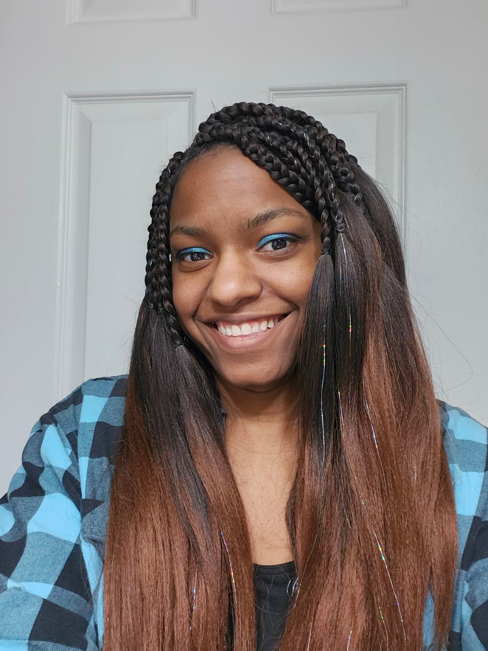 Photograph of Sam Hollis-White smiling while in front of a door. She is wearing a blue and black plaid shirt and is also wearing blue eyeshadow.