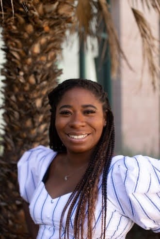 Photograph of Tayjia Wright smiling outside in front of a palm tree. She is wearing a white and blue striped dress. 