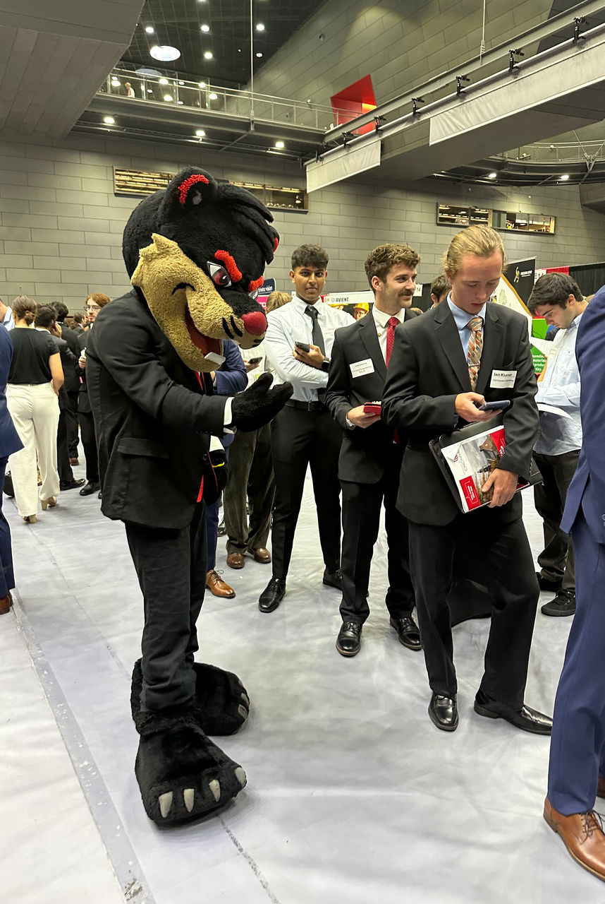 The University of Cincinnati Bearcat mascot, dressed in a black suit and red tie, poses with a group of students in business attire at a busy indoor career fair. The students hold folders and wear nametags, while the background shows other attendees and booths in a large, modern event space.