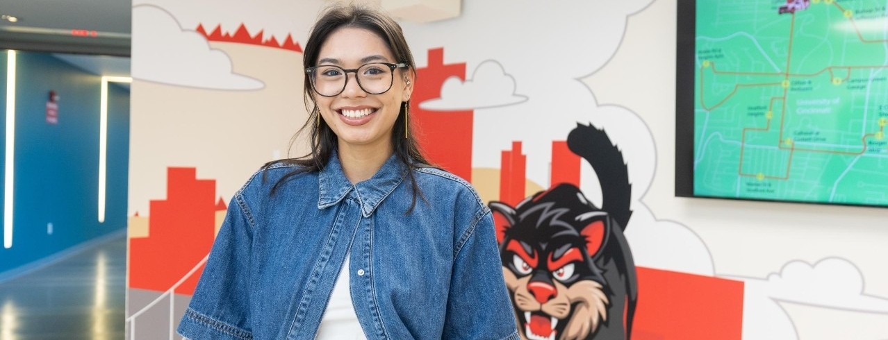 A person smiling in a lobby with "Scioto" and a mascot depicted in the background. The background includes a graphic of a map and city skyline.