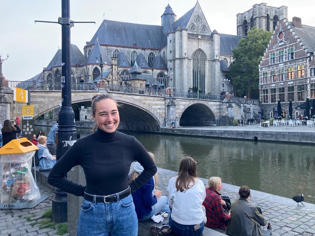 A person smiling in front of the Saint Michael's Bridge with the Church of Saint Michael in the background, in Ghent, Belgium. Several individuals are sitting by the riverbank.