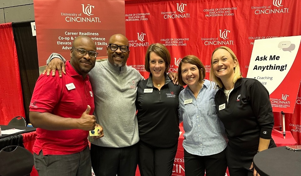 Five people smiling and posing together at a University of Cincinnati event, standing in front of banners with university logos. They wear red and black attire with the university's emblem.