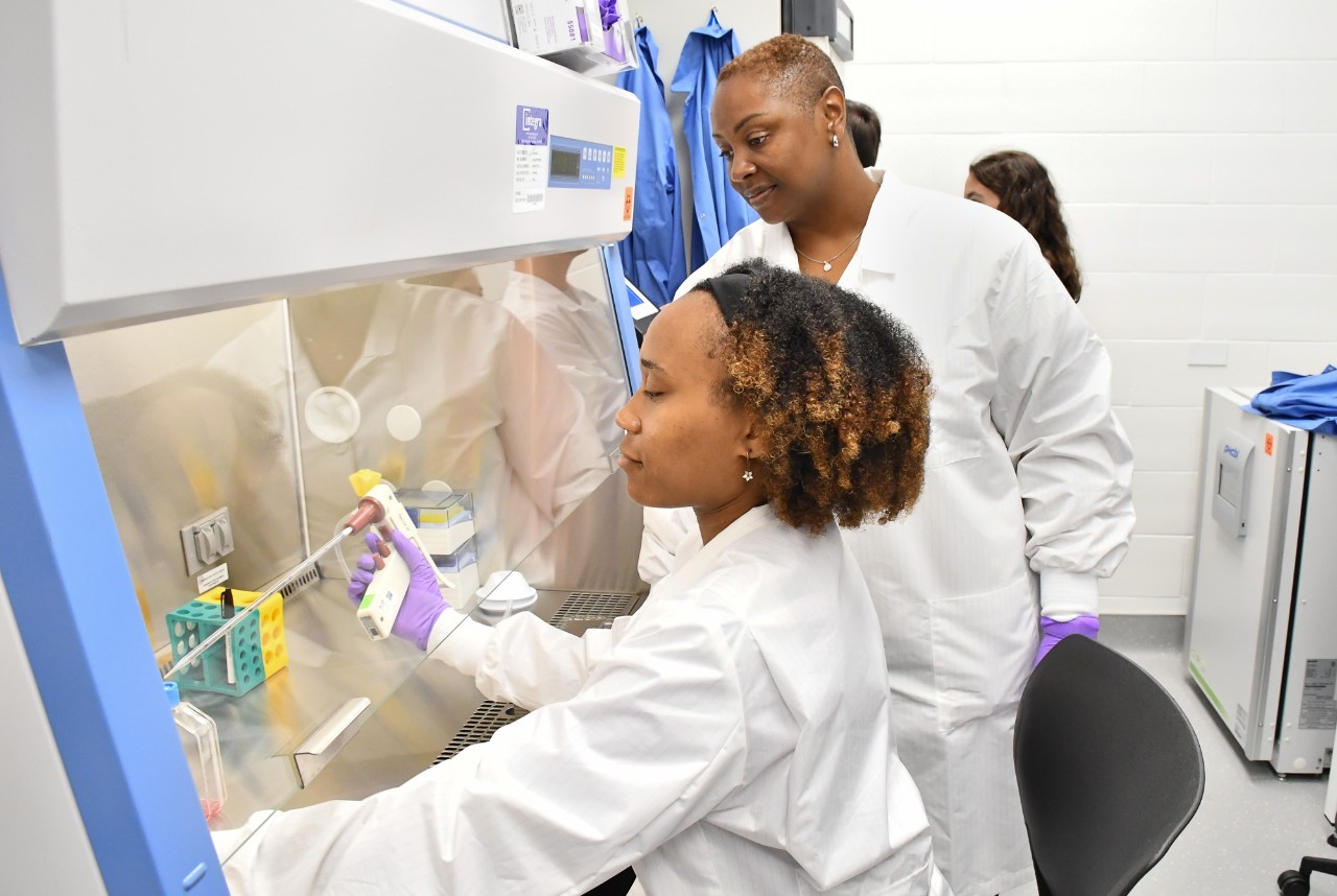 Two scientists working in a laboratory, one using a pipette at a fume hood while the other observes. Both are wearing lab coats.