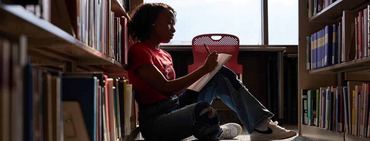 a student sits on the floor in the library between two stacks of books, writing on a notebook propped up on their knee. 