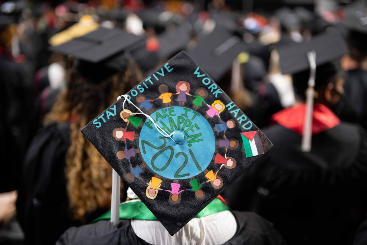 A birds eye view of a graduation cap reading "Stay positive, work hard, 2021" with a drawing of the Earth surrounded by people holding hands.