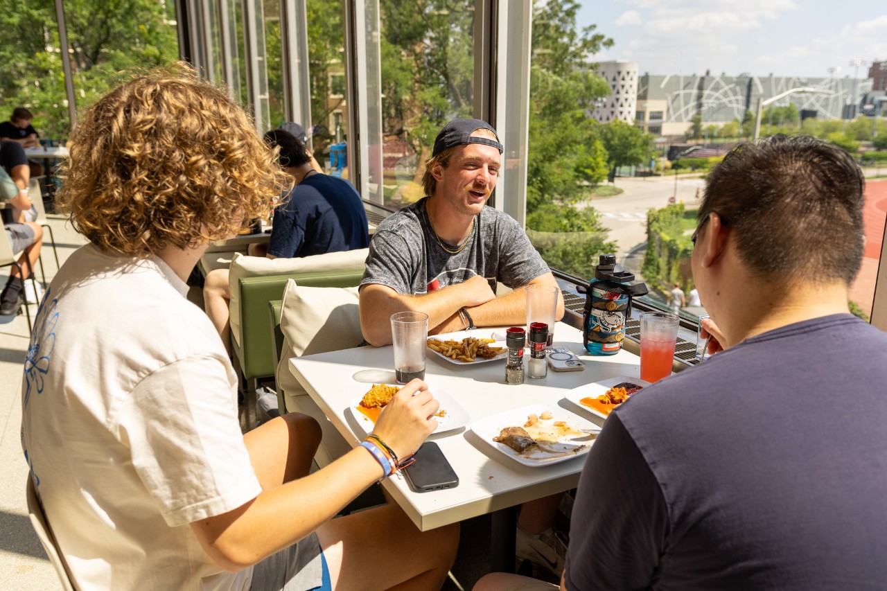 A student smiling while holding a plate of food.  He is holding a black bag that is hanging from his right shoulder. There is a buffet in the background.