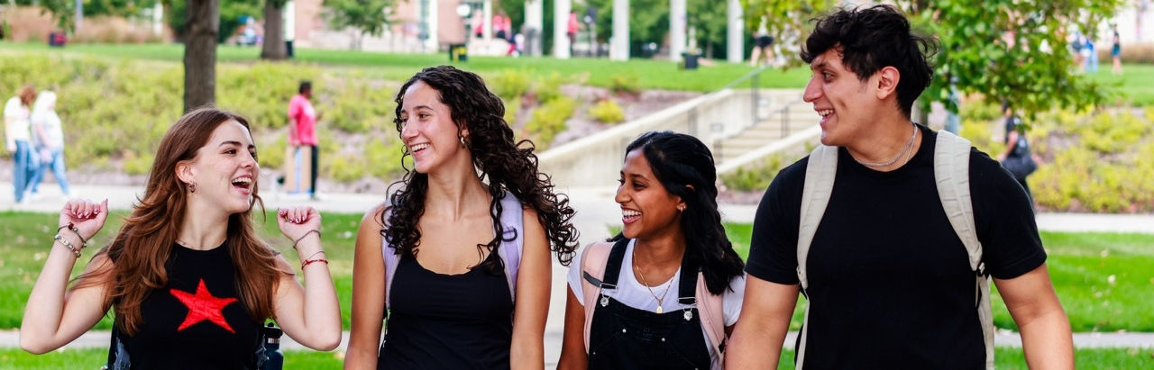 Four UHP students are smiling walking across campus green space. 